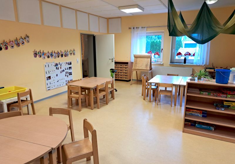 Kita Villa Kunterbunt Titelbild Heller Gruppenraum der Kita Villa Kunterbunt mit kleinen Holztischen und -stühlen, einem Regal mit Spielen, einer Staffelei am Fenster, aufgehängten Kinderarbeiten an der Wand und einem grünen Stoffdach über einem Tisch.