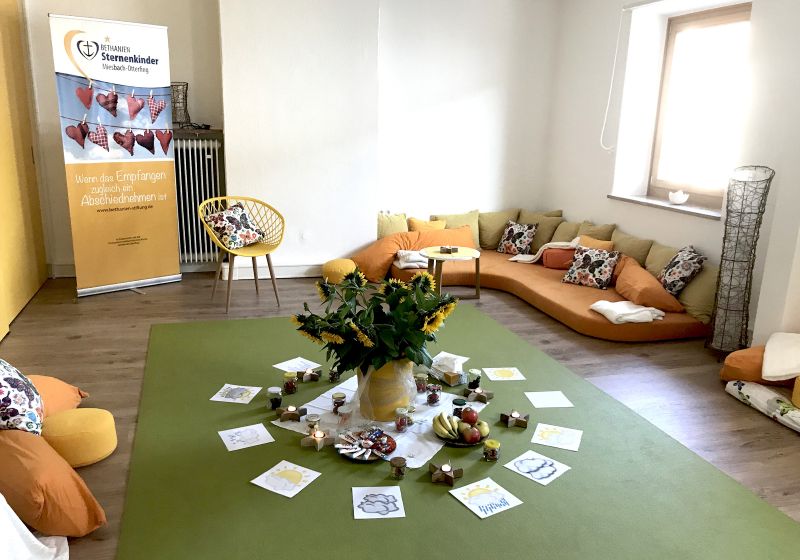 Sternenkinder Oberland/Inntal - Titelbild Ein gemütlichen Raum der Einrichtung mit einem Sofa und vielen Kissen. In der Mitte des Raumes ist ein großer Sonnenblumenstrauß platziert.