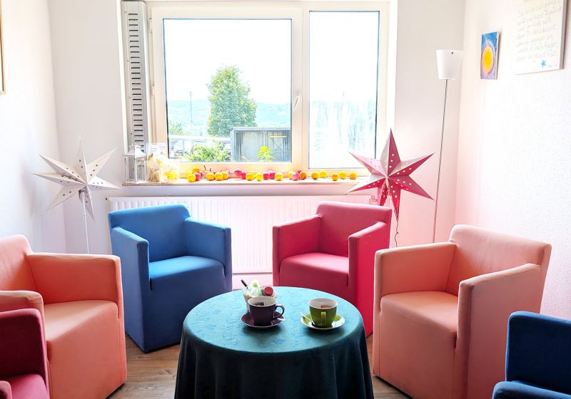 Sternenkinder Wuppertal - Titelbild Das Bild zeigt einen Raum in denen Sessel in unterschiedlichen Farben in einem Kreis stehen. In der Mitte steht ein Tisch mit zwei Tassen und hinter den Sesseln stehen zwei Stern Lampen.